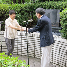 厚労省玄関脇の「誓いの碑」前で要望書を秋葉副厚労相に渡す薬被連代表世話人の花井十伍氏