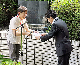 「誓いの碑」前で要望書を田村厚労相に渡す薬被連代表世話人の花井十伍氏(左)