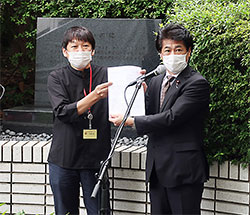 「誓いの碑」前で薬被連代表世話人の花井十伍氏(左)が要望書を田村厚労相に渡した