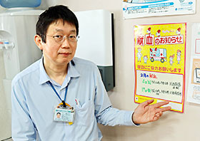 自薬局内に掲示した献血推進のポスターを指す川西市薬剤師会の樋口淳一会長